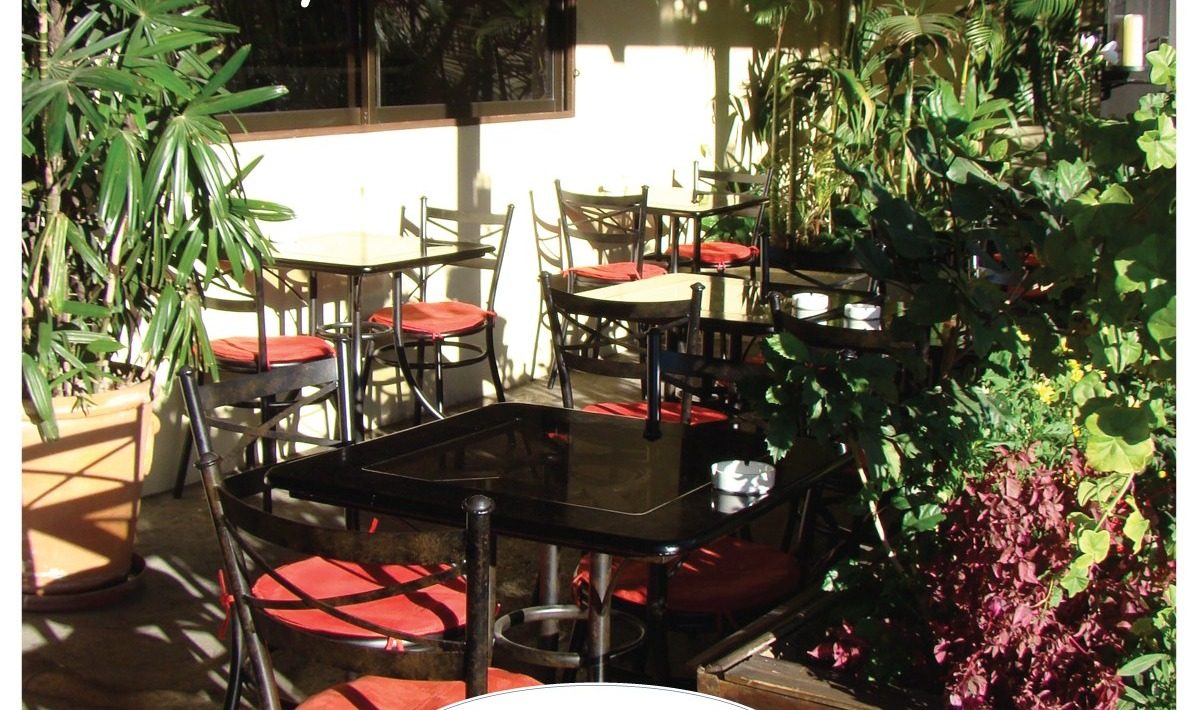 Terraza soleada con mesas negras de bistró, cojines rojos y plantas alrededor